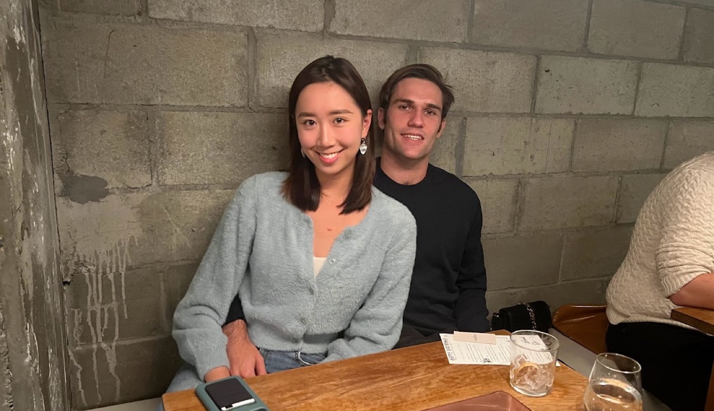 Noel and a woman in a fuzzy light-blue cardigan smile at a wooden restaurant table set against a cinder-block wall.