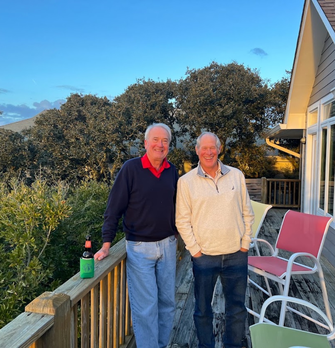 Two older men grin on a weathered beach-house deck at dusk, a bottle in a green monogrammed koozie resting on the railing beside pastel patio chairs.