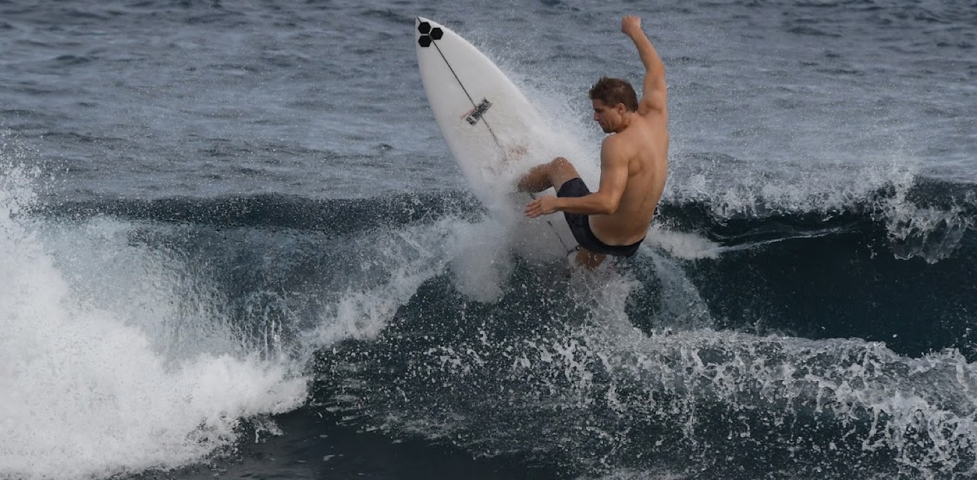 A surfer throws his board vertical off the lip of a churning gray wave, one arm flung overhead in the spray.