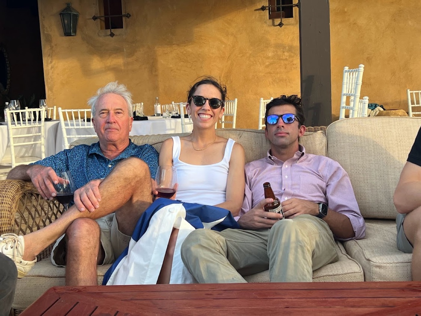 Three guests, an older man holding red wine, a smiling woman in a white tank top, and a man in mirrored sunglasses with a beer, lounge on an outdoor couch against an ochre wall at golden hour with banquet tables behind them.