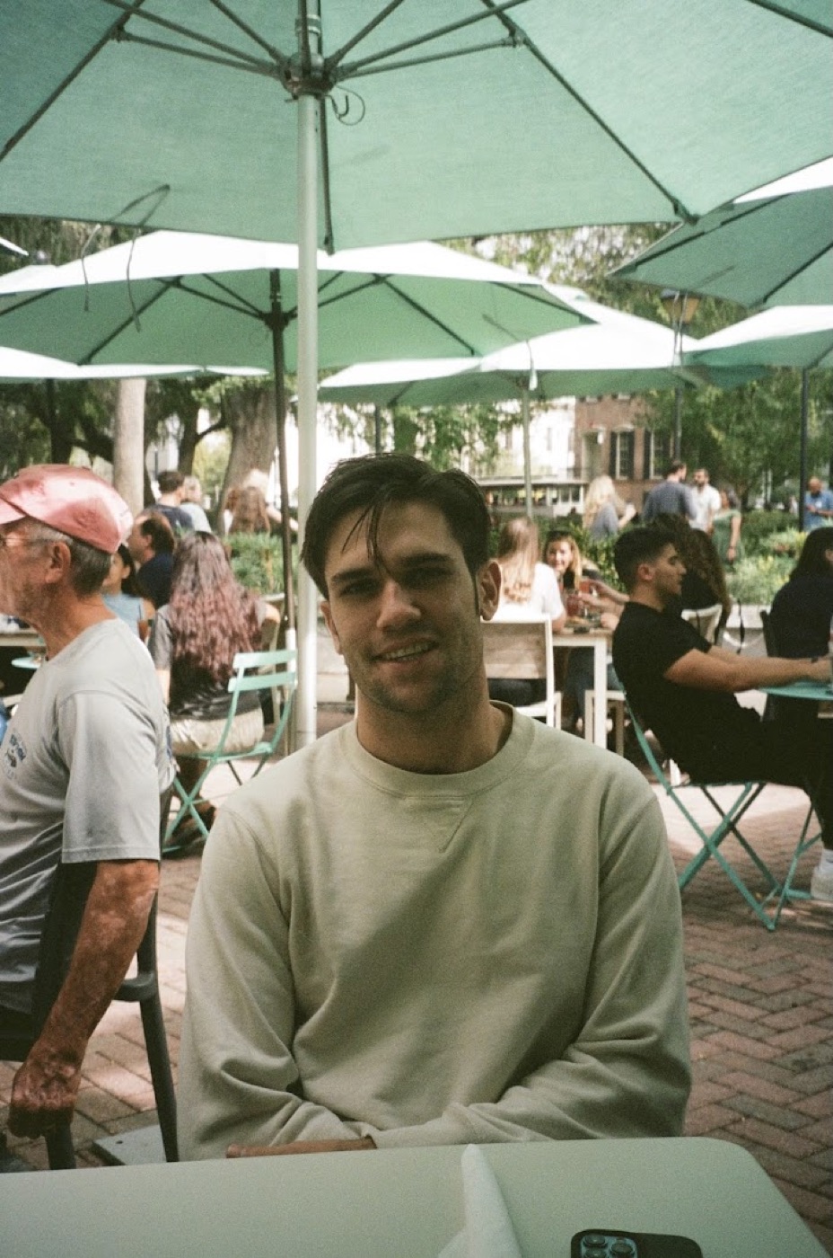 Film photo of Noel in a cream sweatshirt smiling at an outdoor cafe table beneath mint-green umbrellas in a busy tree-lined square.