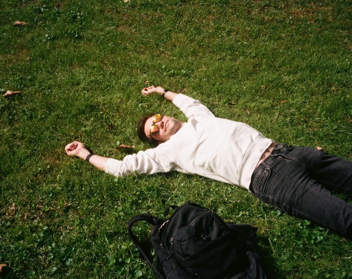 Film photo of Noel sprawled on his back in bright green grass wearing mirrored sunglasses, arms flung wide, a backpack lying at his feet.