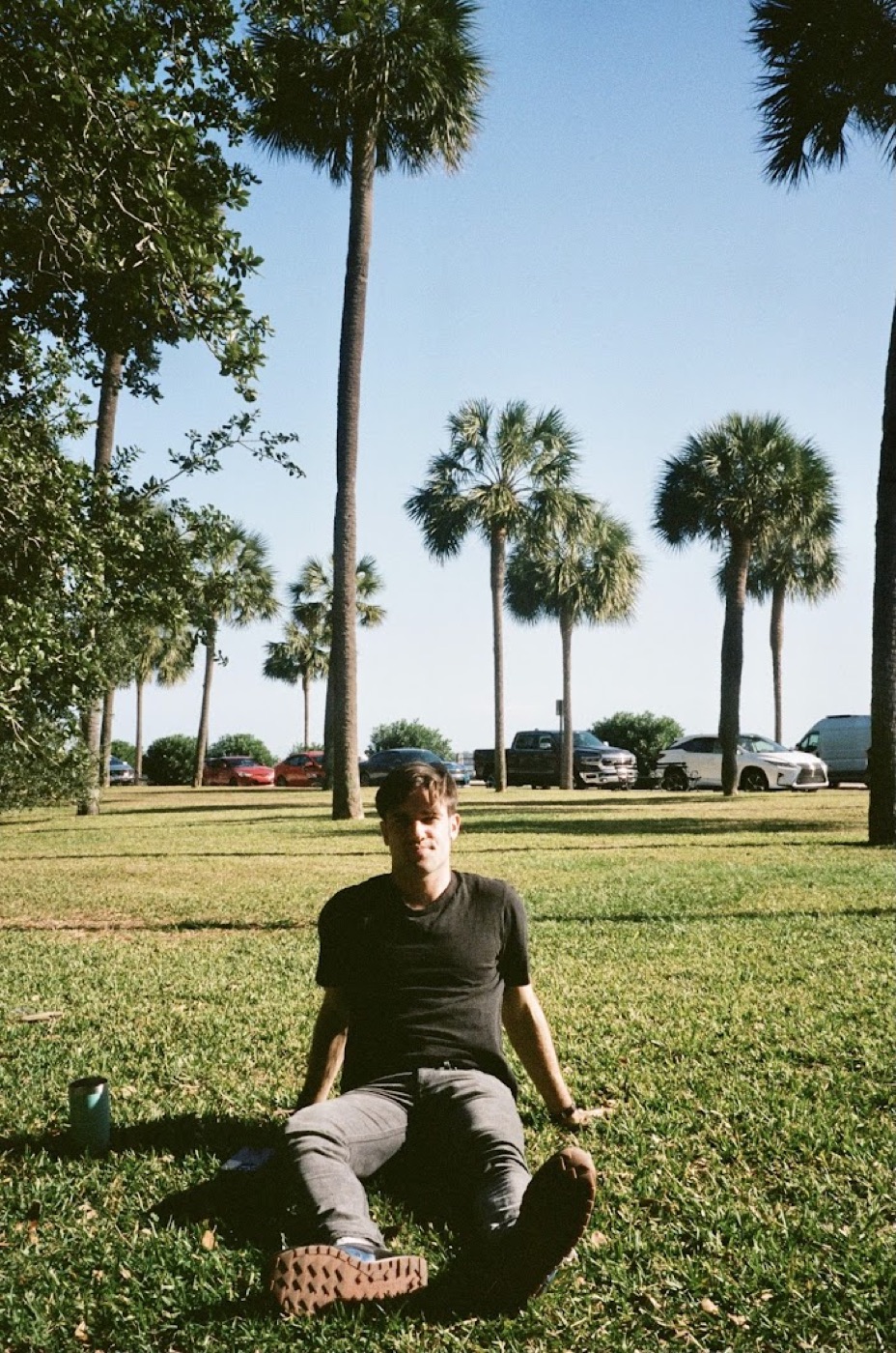 Film photo of Noel sitting on a lawn in a black t-shirt with a canned drink beside him, tall palm trees and parked cars in the background.