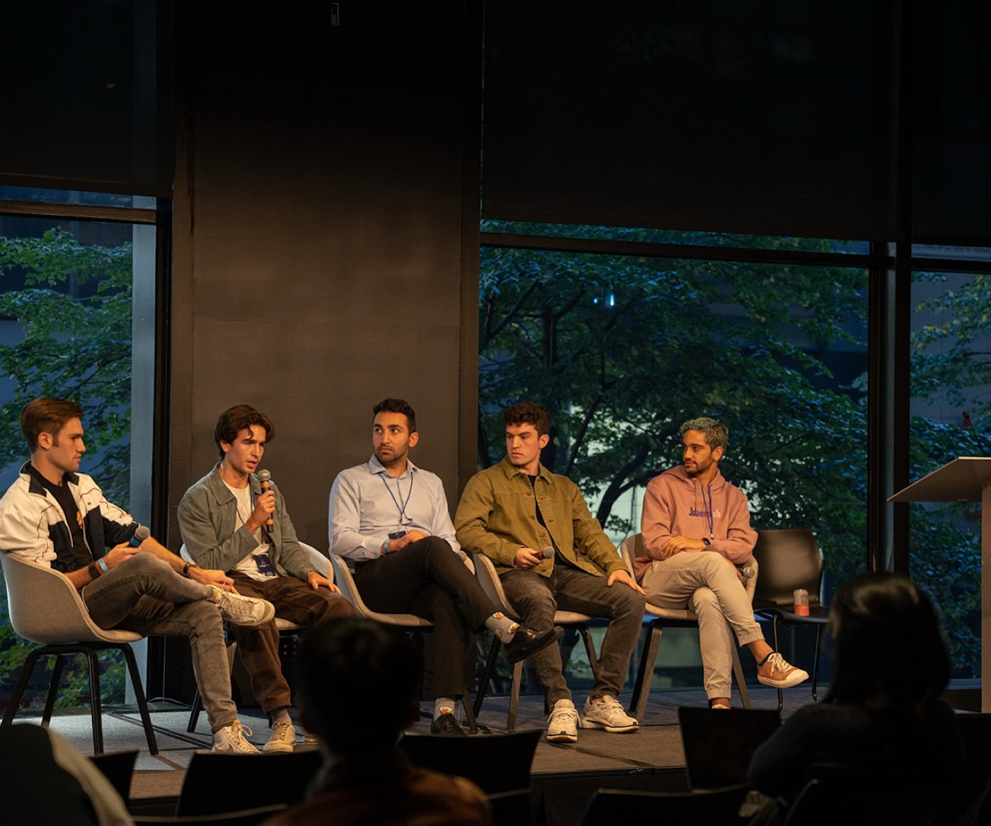 Noel, in a retro white-and-navy track jacket, listens from the end of a five-person panel while another speaker talks into a microphone in front of tall windows.