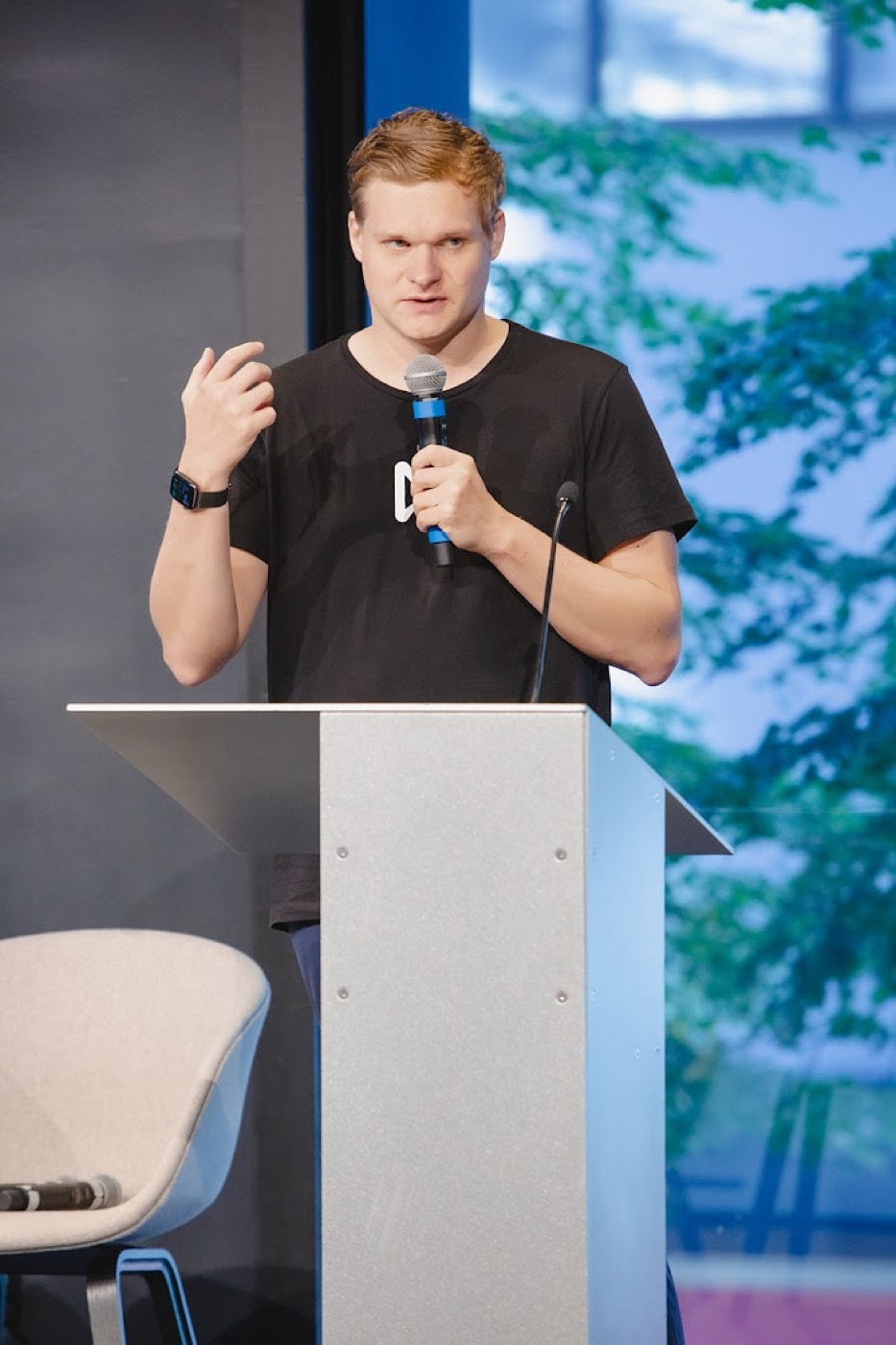 A red-haired man in a black t-shirt gestures while speaking into a handheld microphone from behind a silver podium.