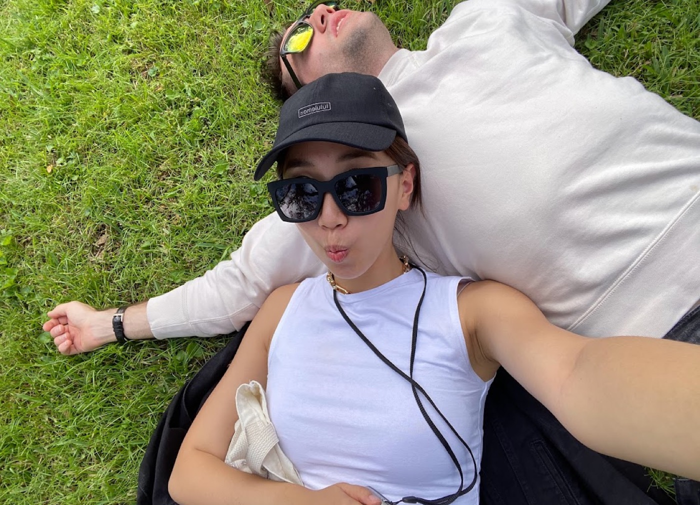 Overhead selfie of a woman in a black cap and oversized square sunglasses lying head-to-head in the grass with Noel, who stretches out behind her in a white sweatshirt.
