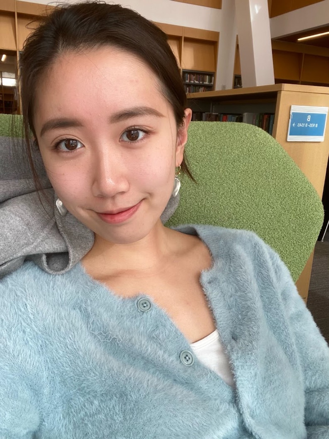 Close selfie of a smiling woman in a fuzzy blue cardigan settled into a green armchair between library bookshelves.