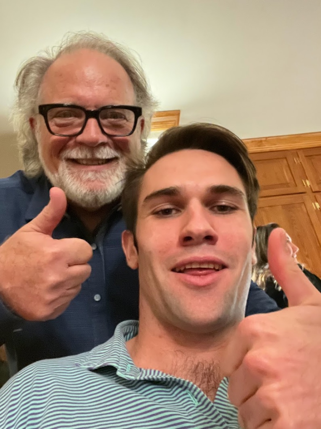 Selfie of Noel and a white-bearded older man in thick black glasses, both giving big thumbs up indoors.