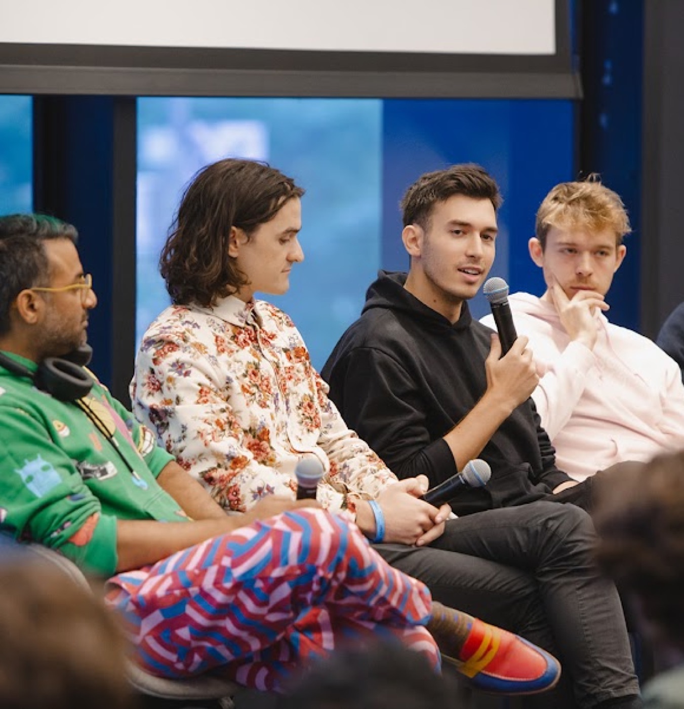 Four panelists sit in a row at an indoor event, one in a floral jacket looking on as a man in a black hoodie answers into a microphone.
