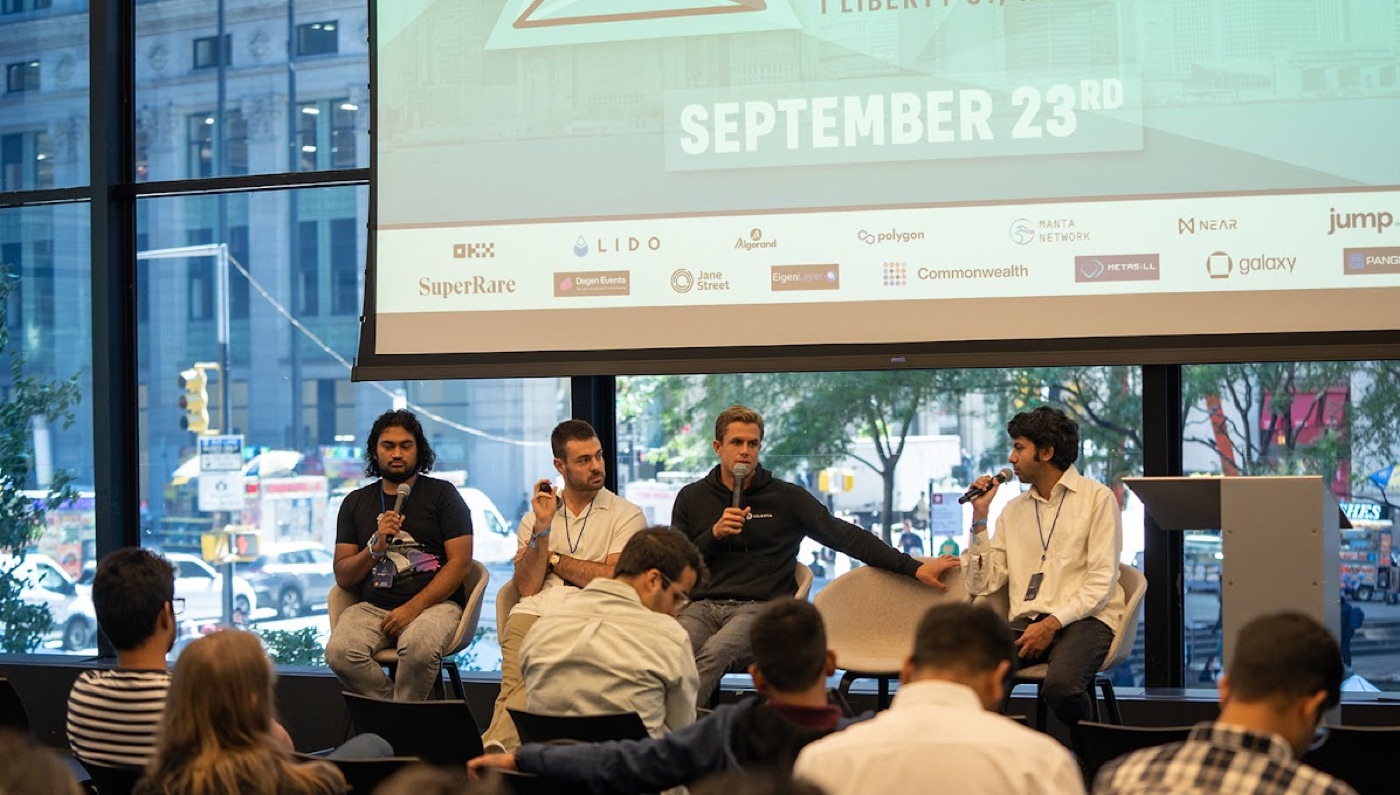 Four speakers hold microphones on a conference stage beneath a projection screen dated September 23rd and rows of crypto sponsor logos.