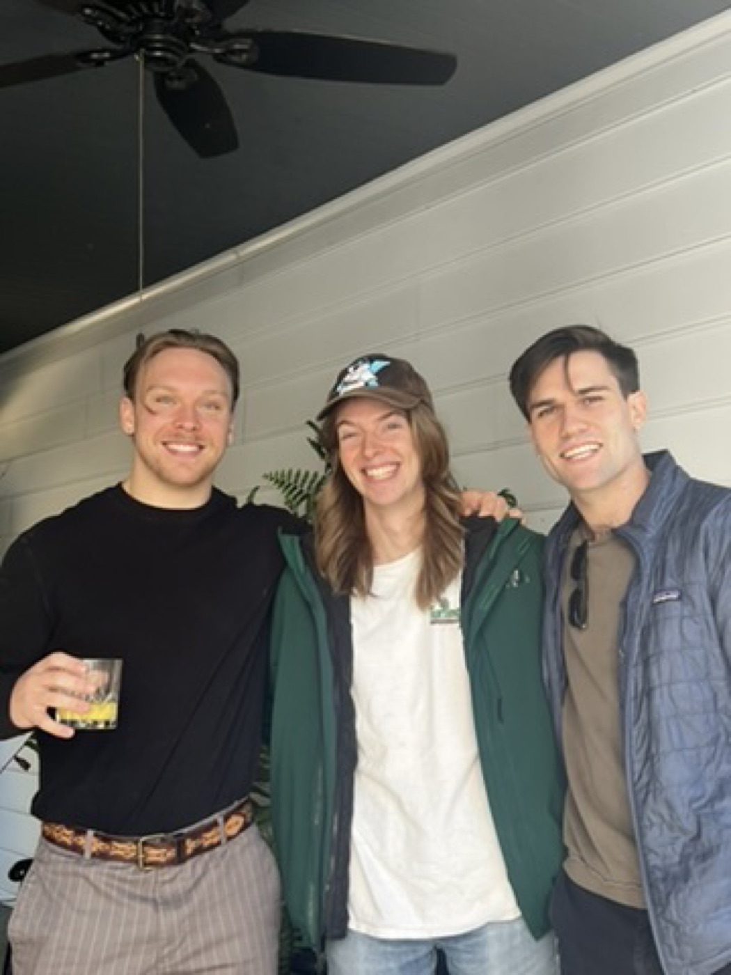Three friends grinning arm-in-arm on a cozy porch, drinks in hand