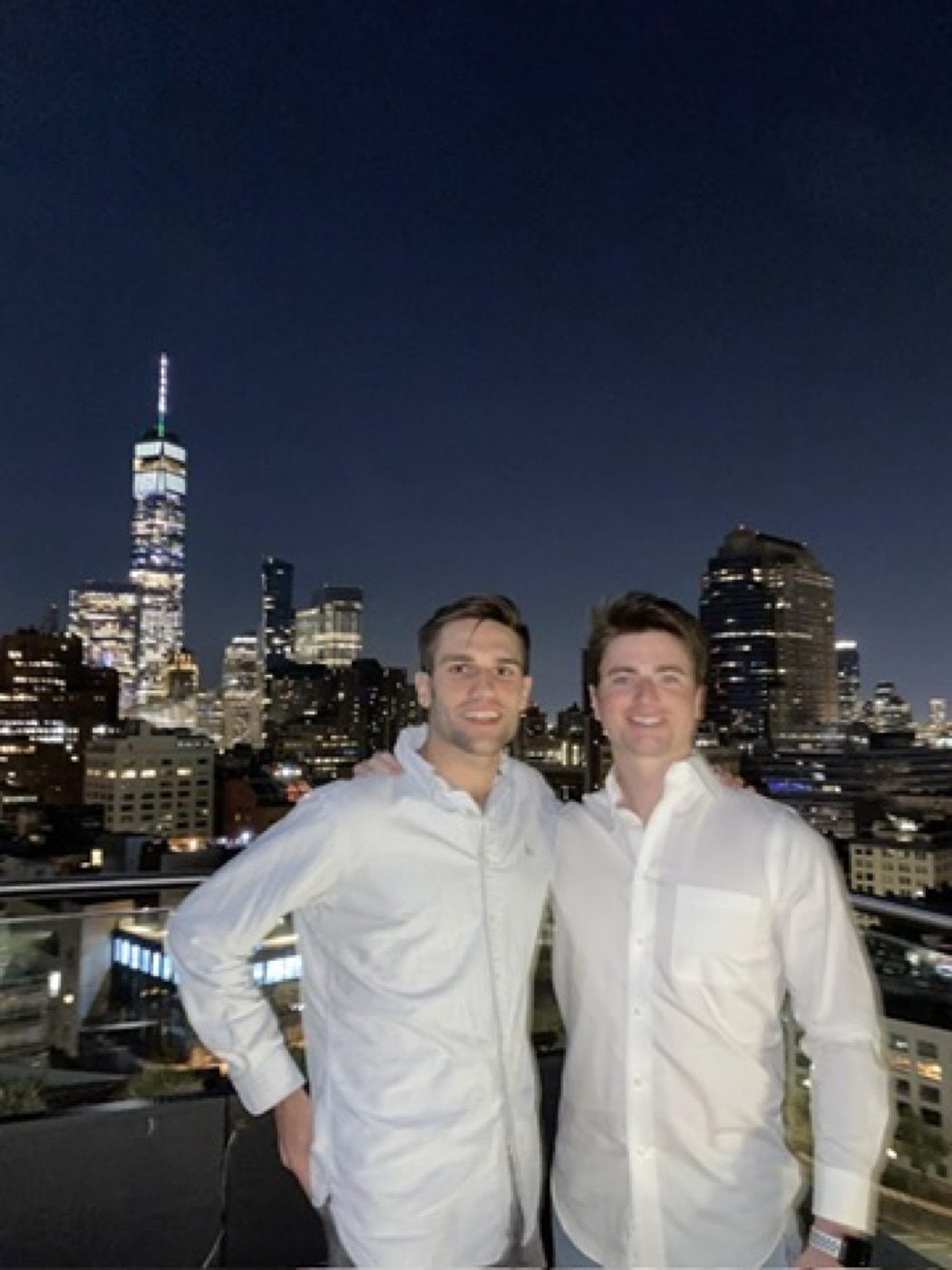 Two men in white shirts before the glowing Manhattan skyline at night