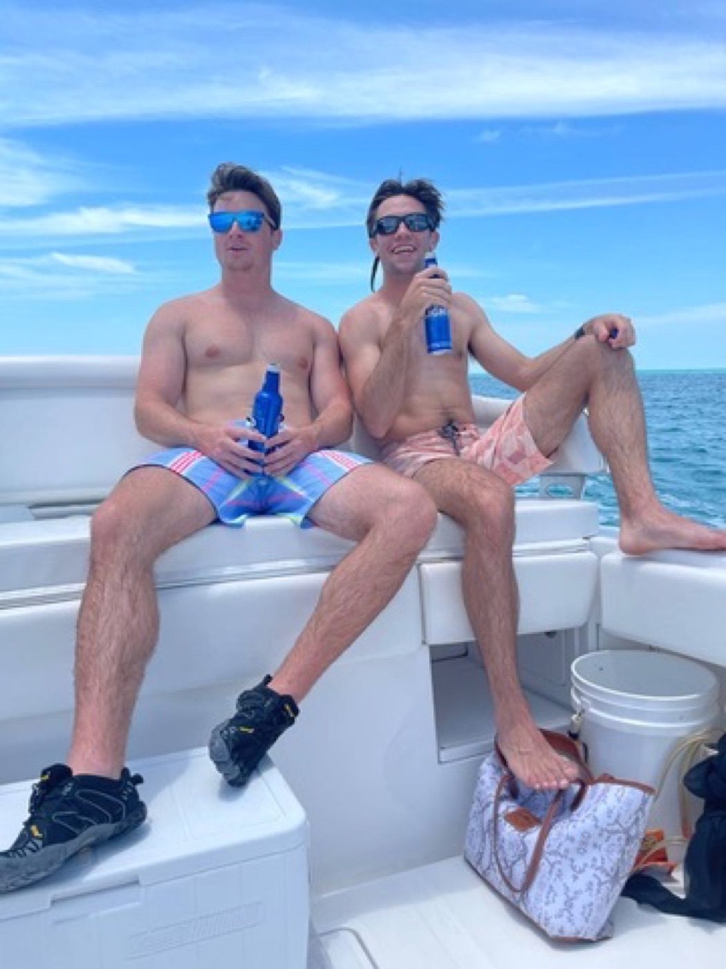 Two shirtless friends relaxing with drinks on a boat over teal ocean