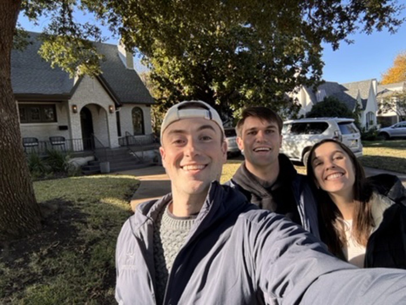 Arms-out selfie with his partner and friend by an autumn house