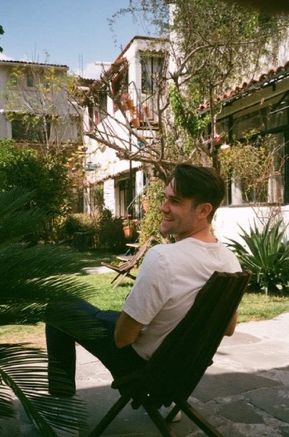 Noel laughing in a chair in a sunny green courtyard