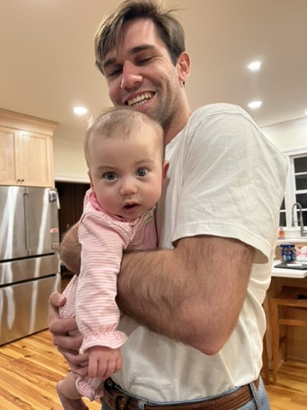 Grinning while cradling a wide-eyed baby in the kitchen