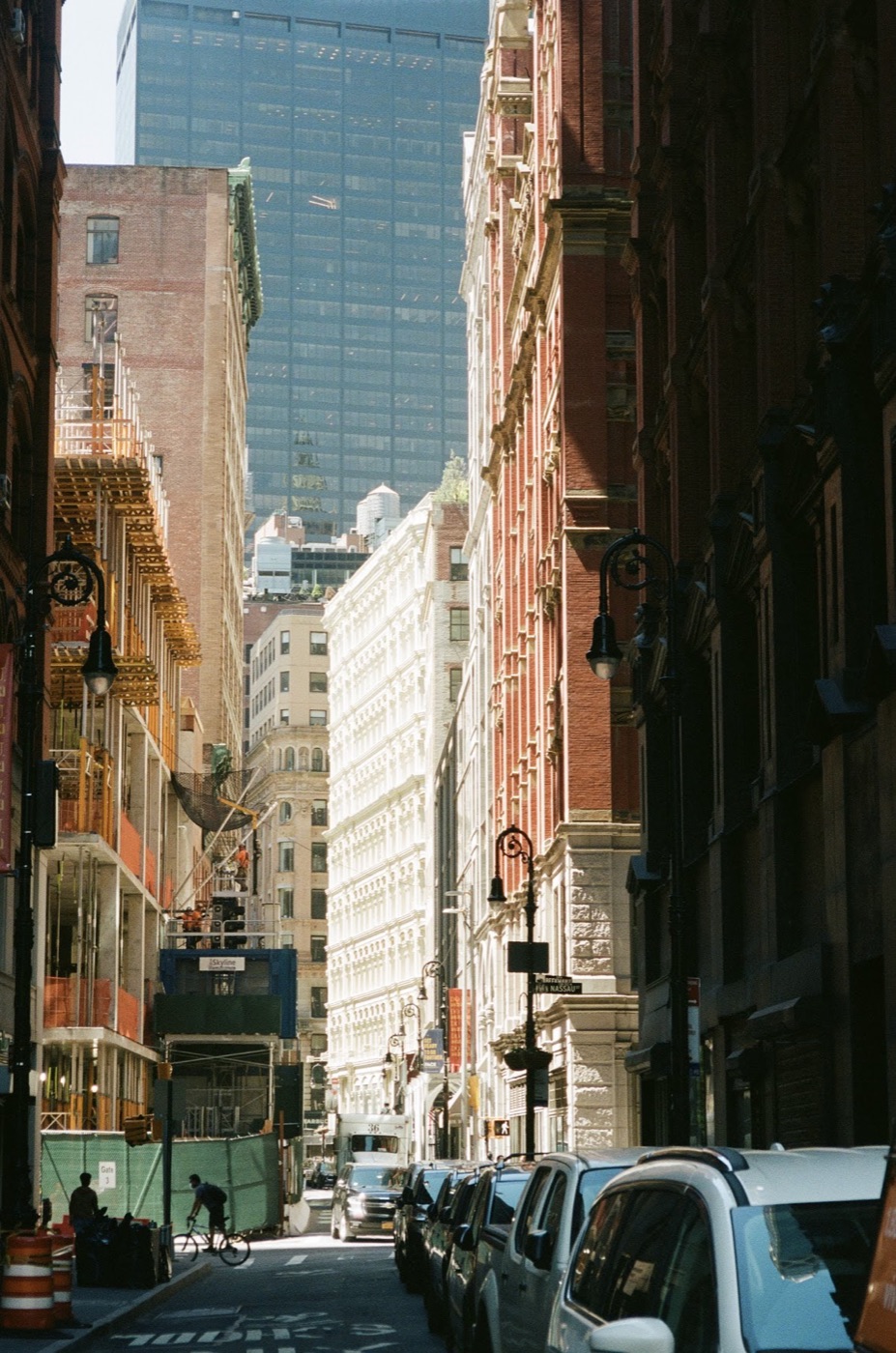Sunlit film photo down a narrow Manhattan street, ornate stone and brick facades and construction scaffolding framing a lone cyclist crossing in the shade.