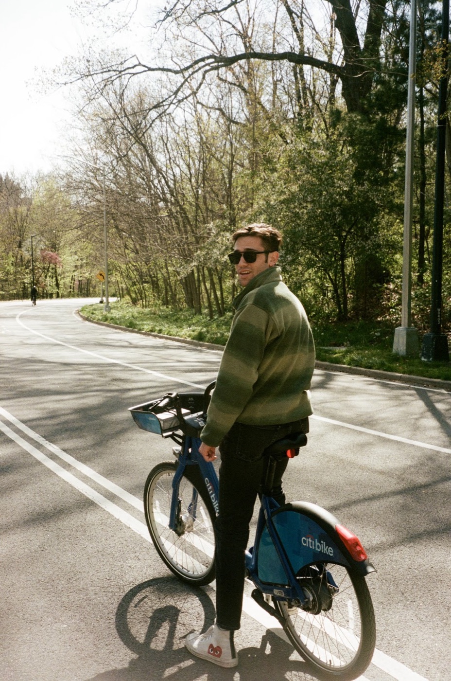 Noel pauses on a blue Citi Bike on a curving park road, looking back over his shoulder in sunglasses and a striped green fleece.