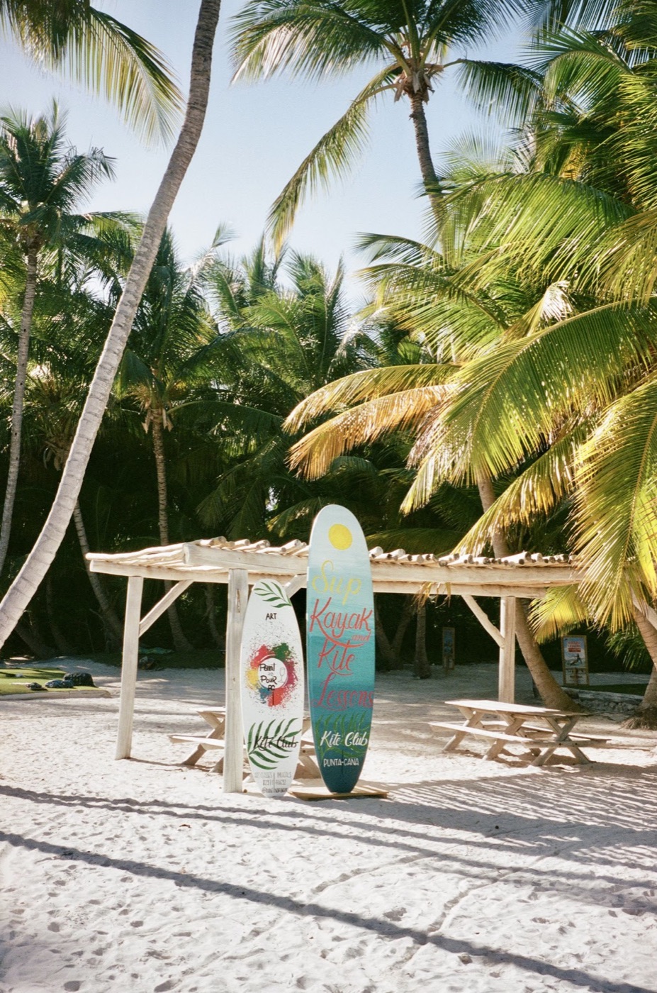 Two hand-painted boards advertising SUP, kayak, and kite lessons stand under palm trees on a white-sand beach in Punta Cana.