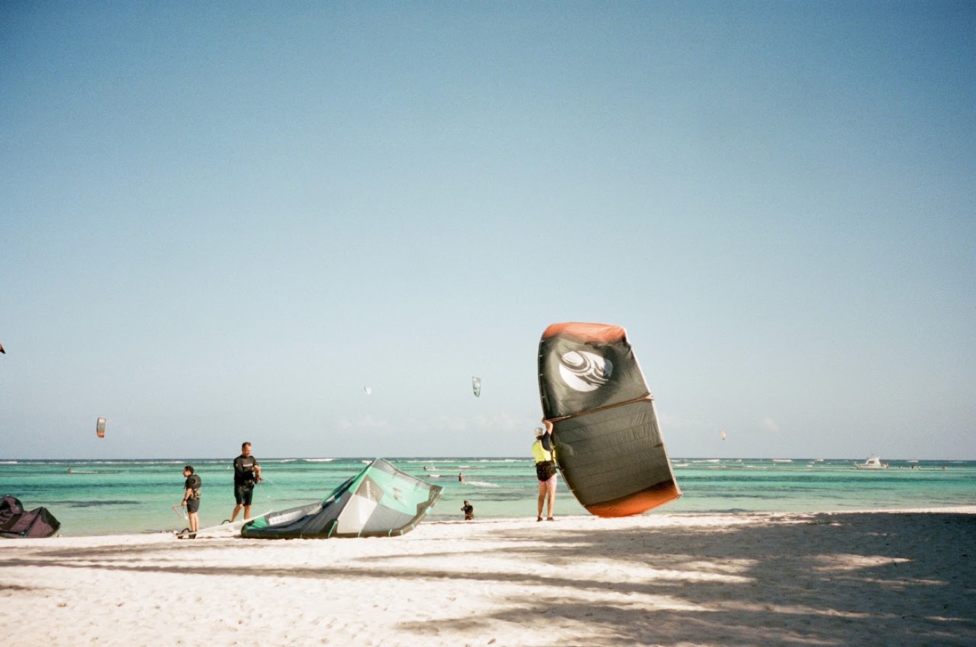 Kiteboarders rig their gear on a white-sand beach beside shallow turquoise water, one hoisting a dark kite overhead.