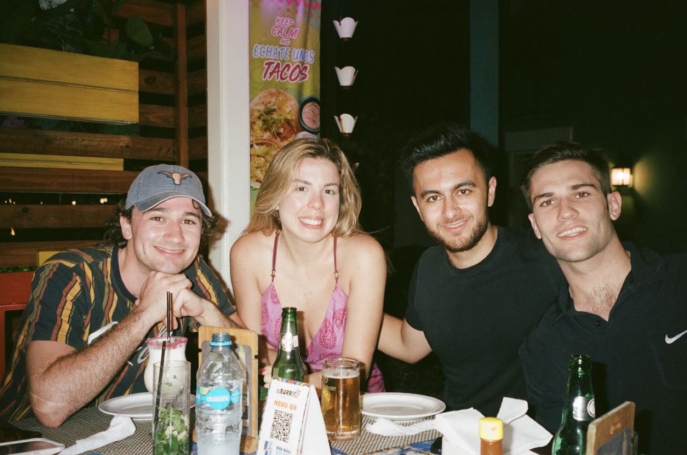Noel and three friends lean together over a taco-restaurant table at night, mojito, beers, and a QR menu card between them.