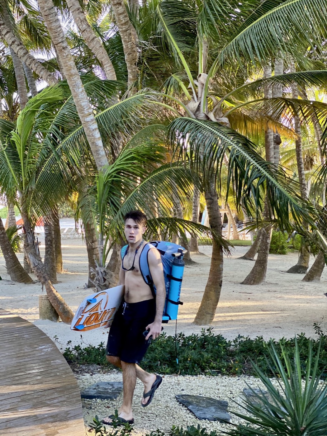 Noel, in board shorts and flip-flops, carries a board under one arm and a blue gear bag on his back through a grove of palm trees.