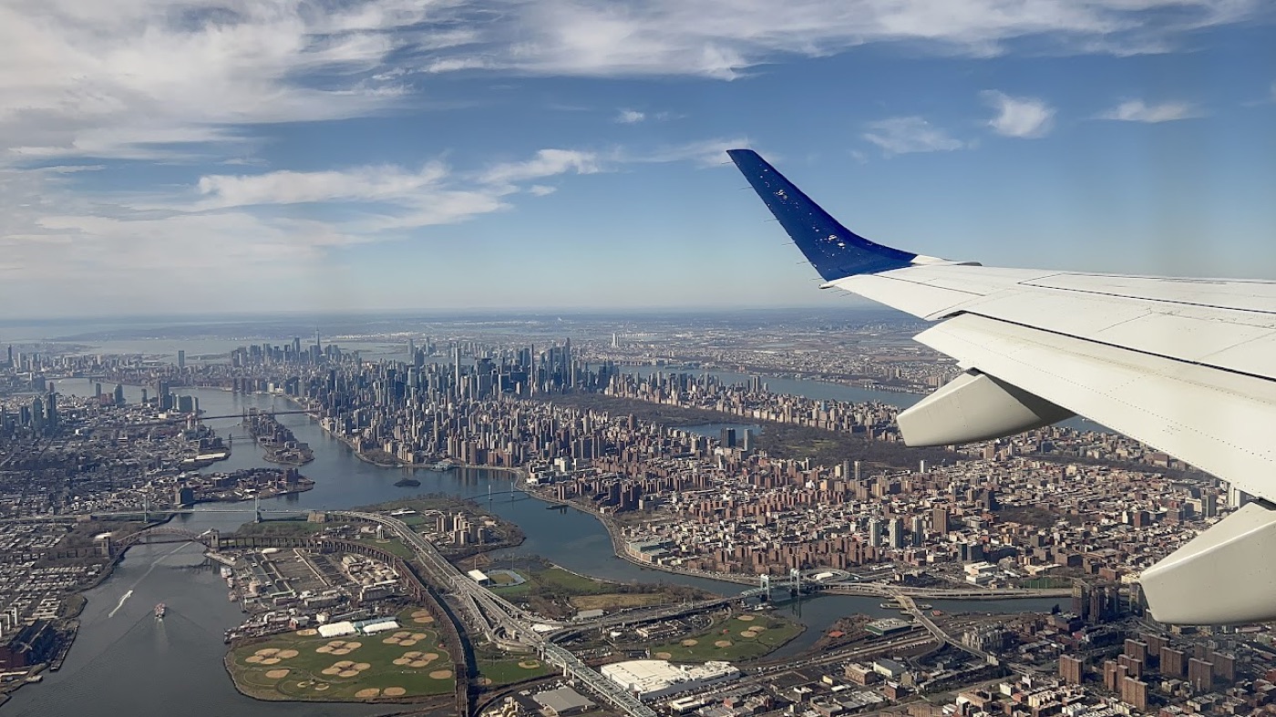 Airplane-window view over the wing of Manhattan and the East River, midtown towers and Central Park hazy in the distance.