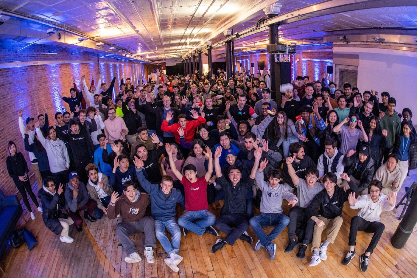 A packed hackathon crowd cheers for a fisheye group photo in a brick-walled loft washed in blue and purple light.