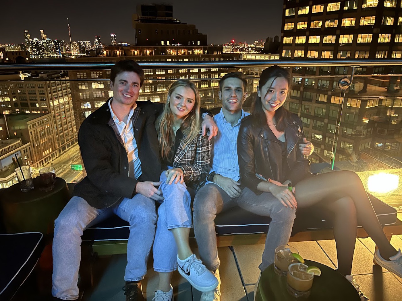 Noel and three friends squeeze onto a rooftop-bar bench at night, cocktails at hand and lit office windows glowing behind them.