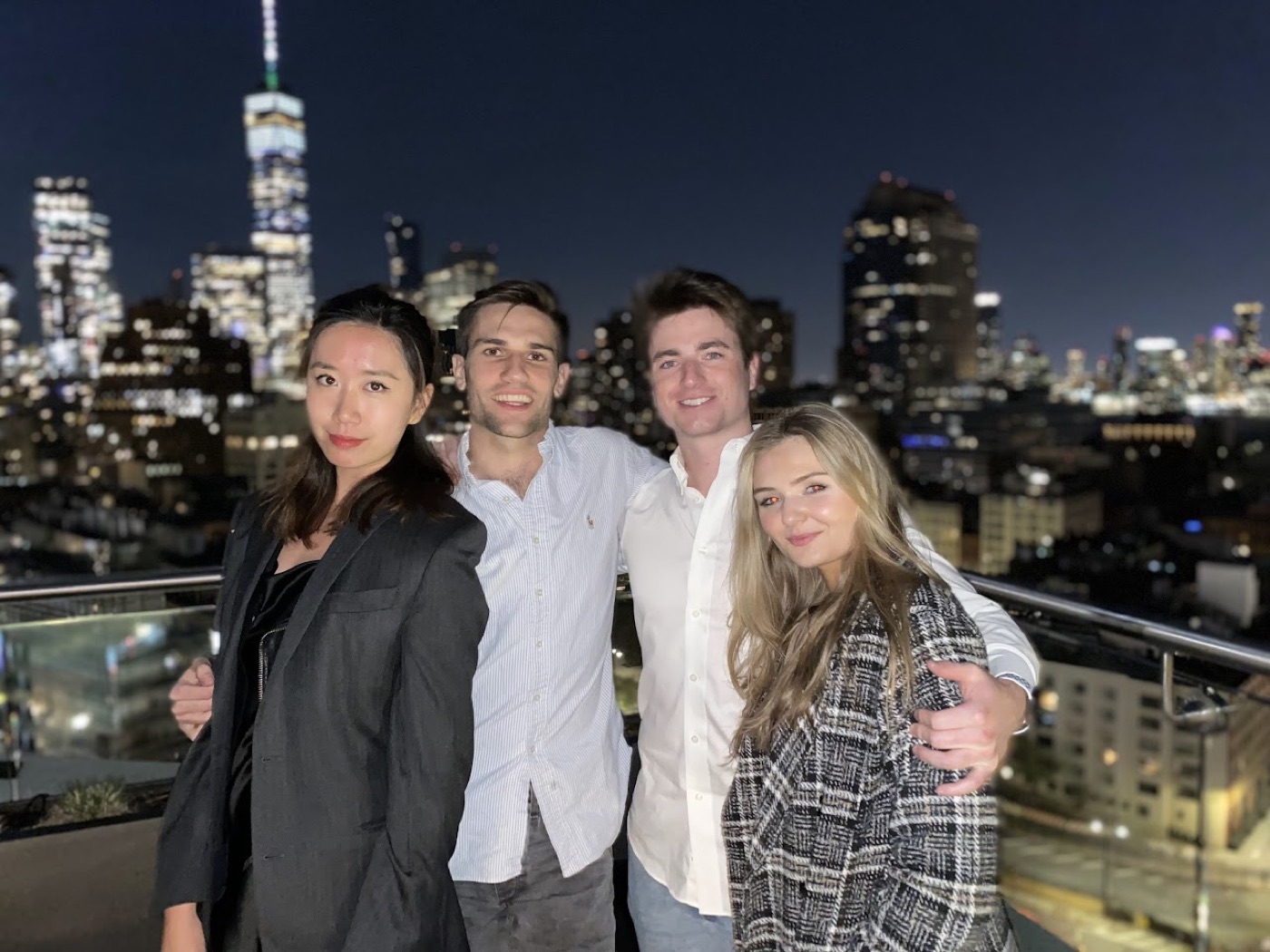 Noel and three friends pose on a rooftop terrace at night with the downtown skyline and One World Trade Center behind them.