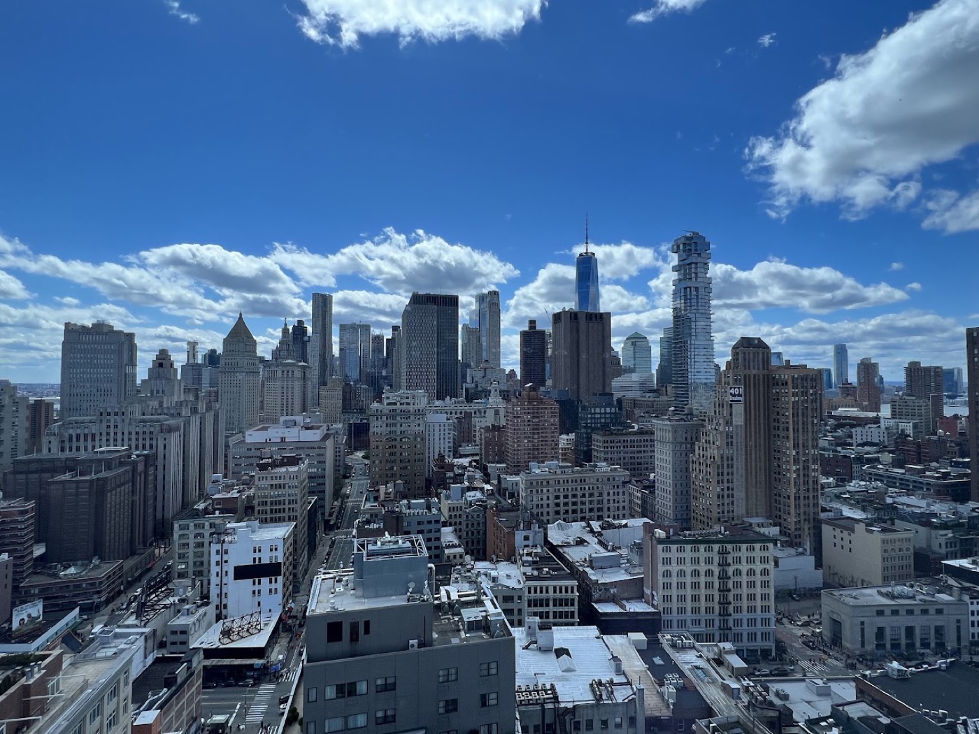 Daytime view from a high floor across lower Manhattan rooftops, One World Trade Center rising into a blue, cloud-scattered sky.