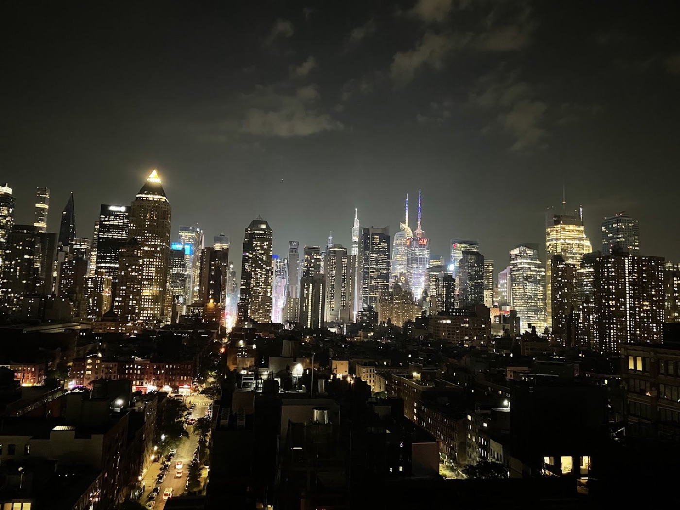 The midtown Manhattan skyline glowing at night, seen from a rooftop over dark tenement roofs and a single lit street.