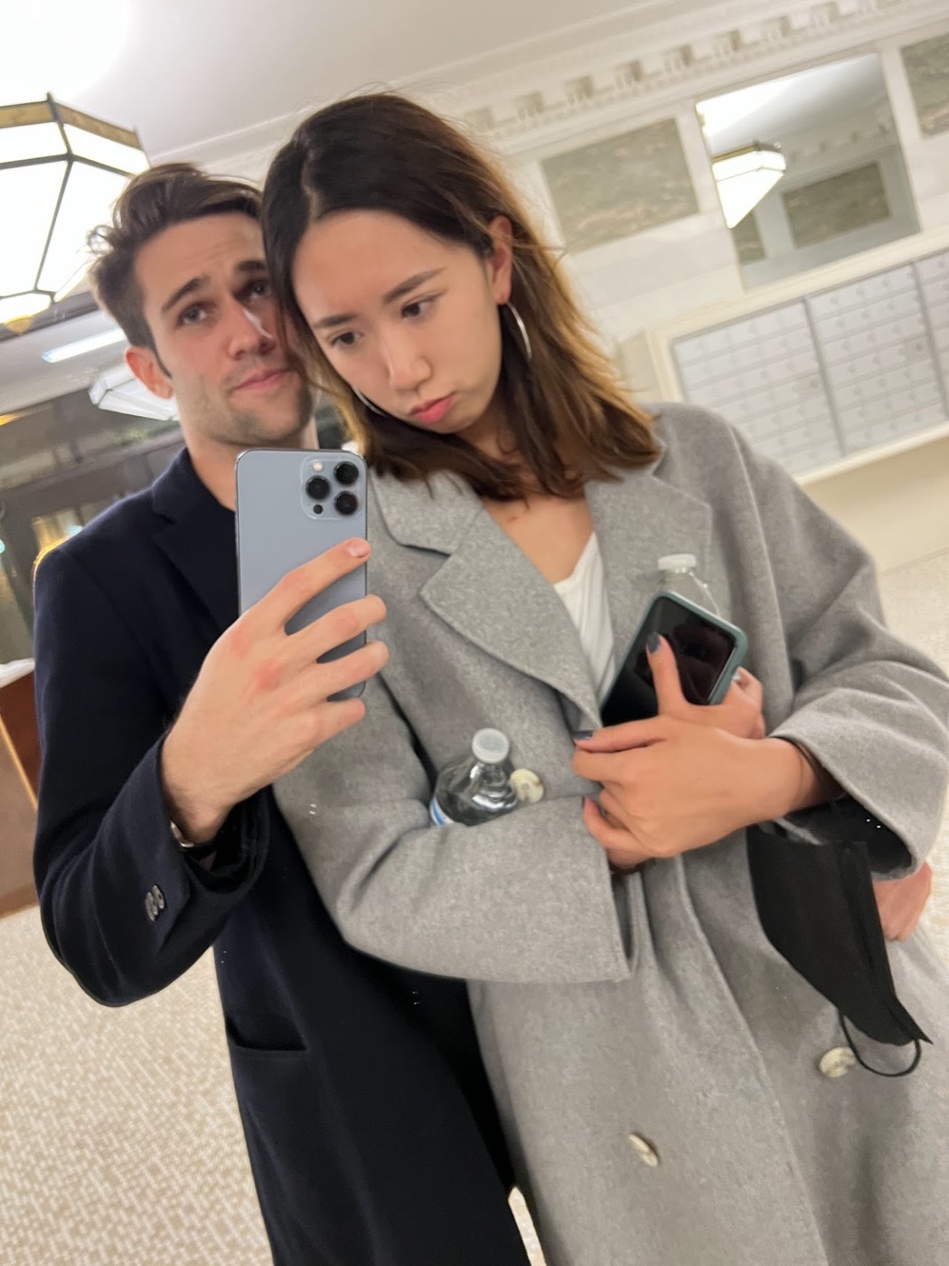 Noel and a companion take a mirror selfie in an apartment lobby, bundled in wool coats with a bank of brass mailboxes behind them.