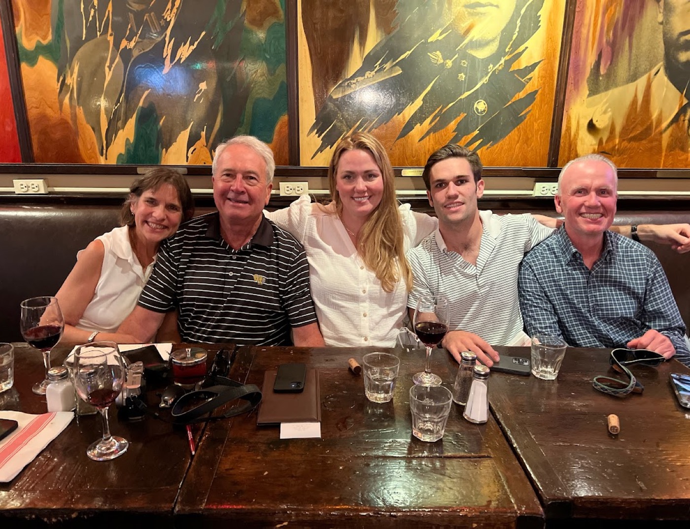 Noel sits among four smiling dinner companions at a scarred wooden restaurant table set with red wine, beneath large torn-portrait paintings.