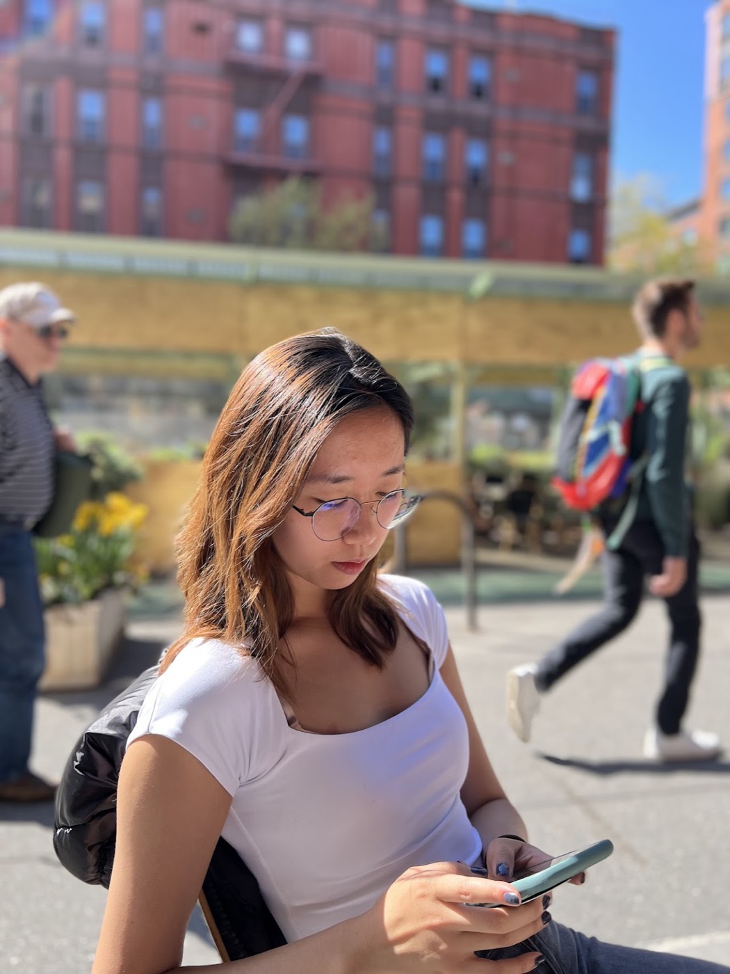 A woman in wire-rim glasses reads her phone at a sunny sidewalk table, a red-brick building and passersby soft-focus behind her.