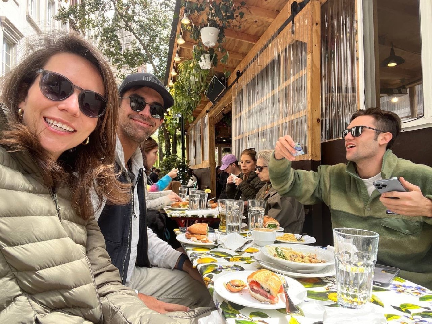 Selfie at a plant-draped outdoor restaurant: a smiling woman in a puffer, a man in a Delta cap, and Noel across the table holding up a card.