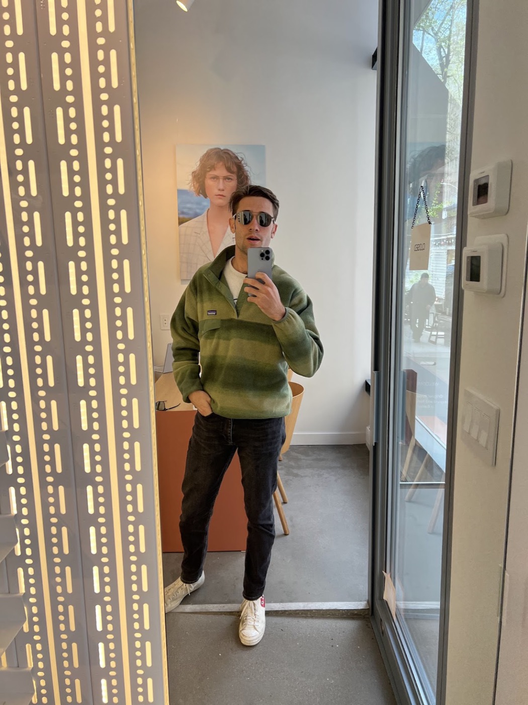 Noel takes a mirror selfie in sunglasses and his striped green fleece inside a small shop, a portrait poster on the wall behind him.