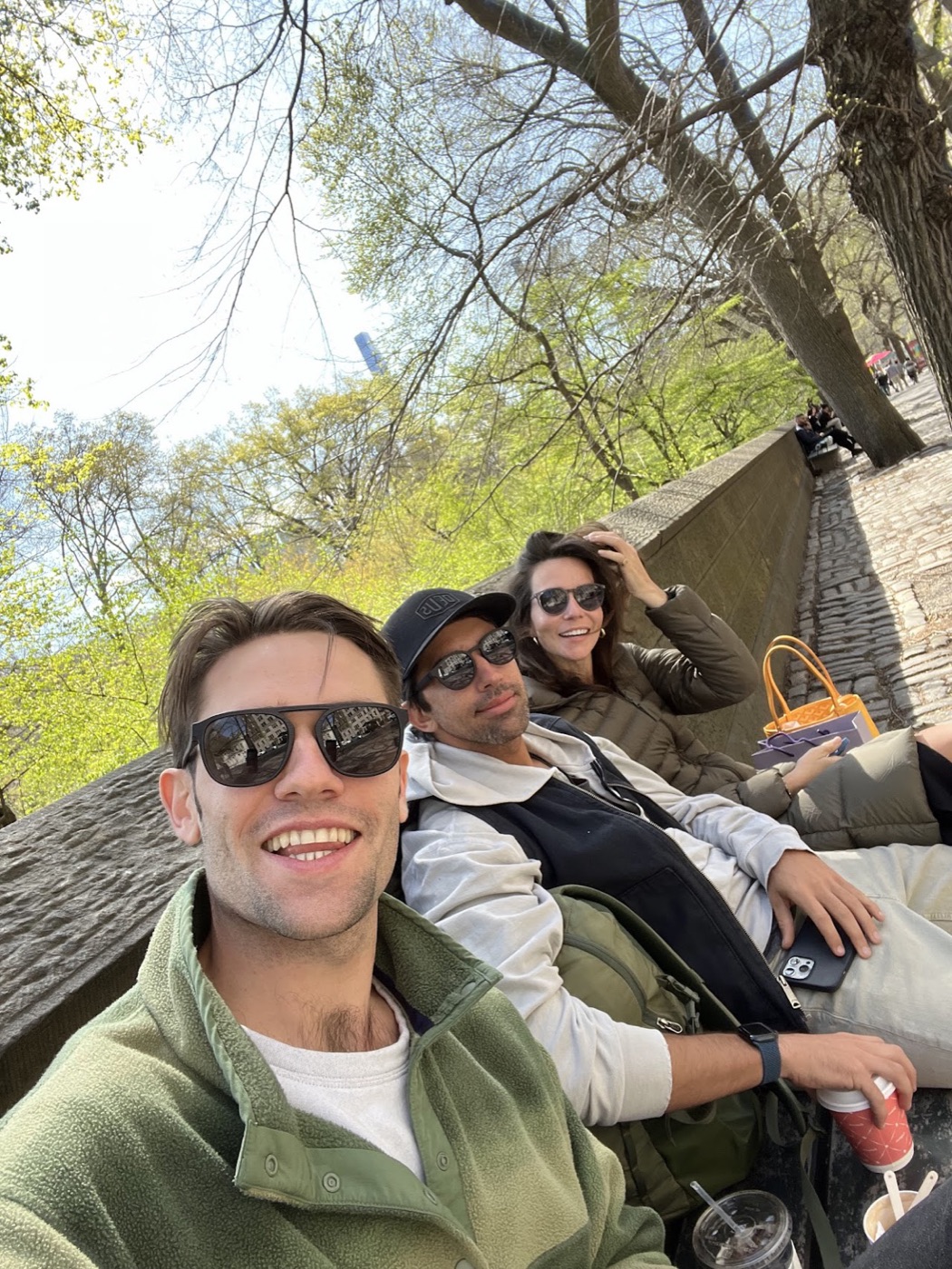 Noel grins into a selfie from a park bench with two friends lounging beside him, bare spring trees and a cobblestone path behind.