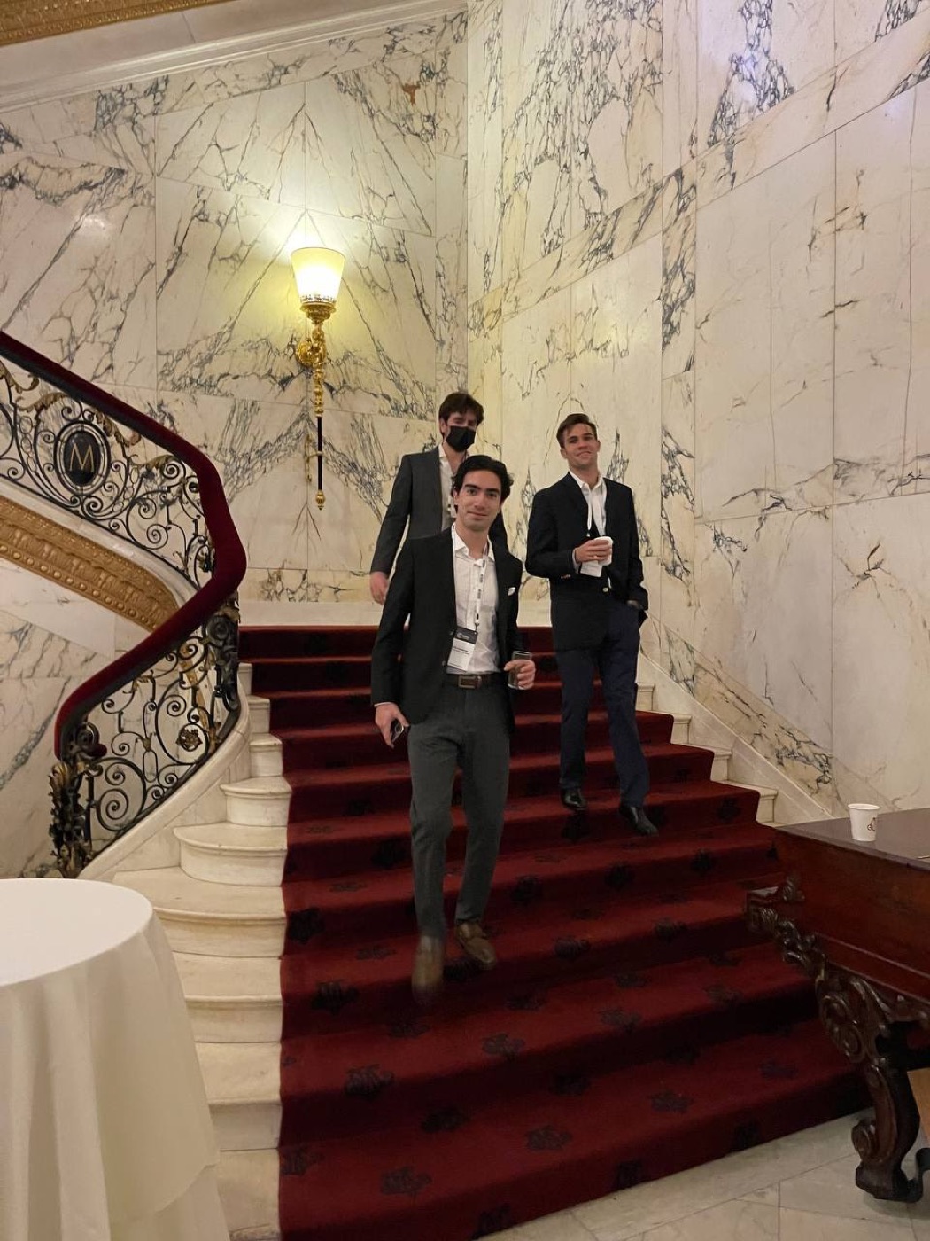 Three young men in blazers and event lanyards descend a red-carpeted marble staircase beneath a gilded sconce.