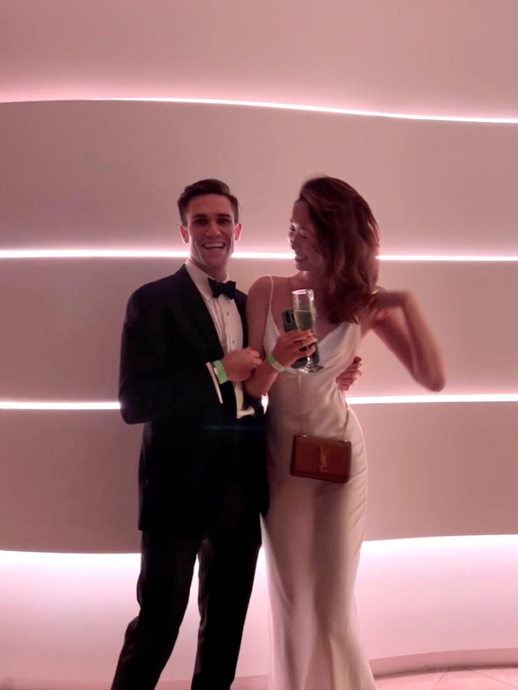 Noel, in a tuxedo, laughs mid-toast with a woman in a white slip dress holding champagne against a wavy wall glowing pink.