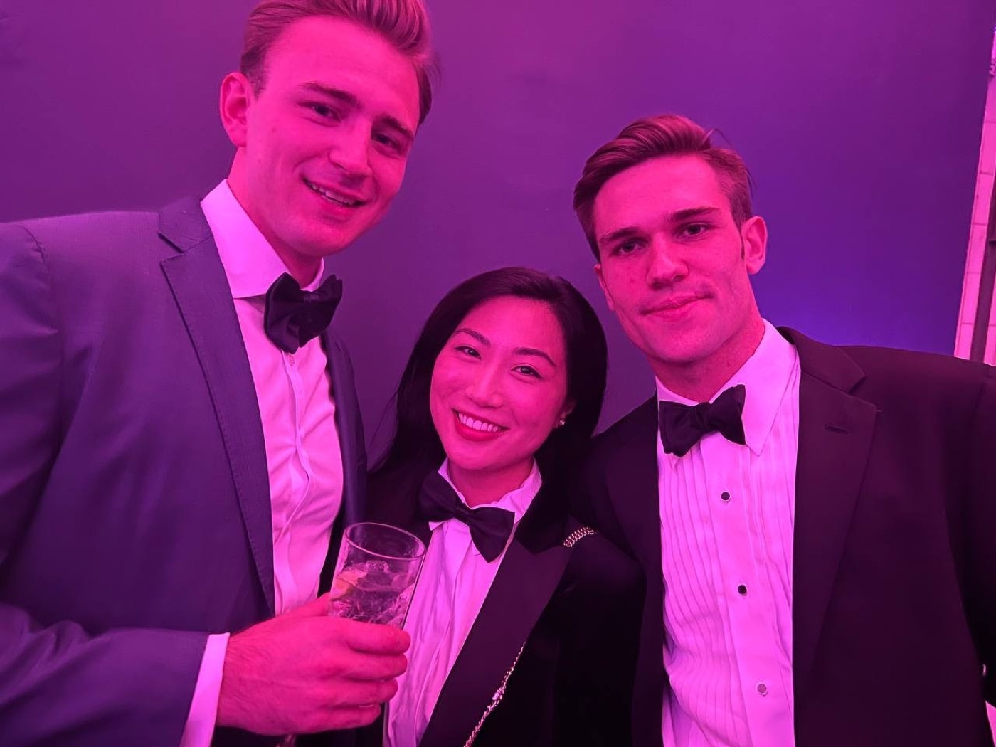 Three friends in black tie, one in a gray suit and a woman wearing a bow tie, crowd into a selfie under magenta party lighting.