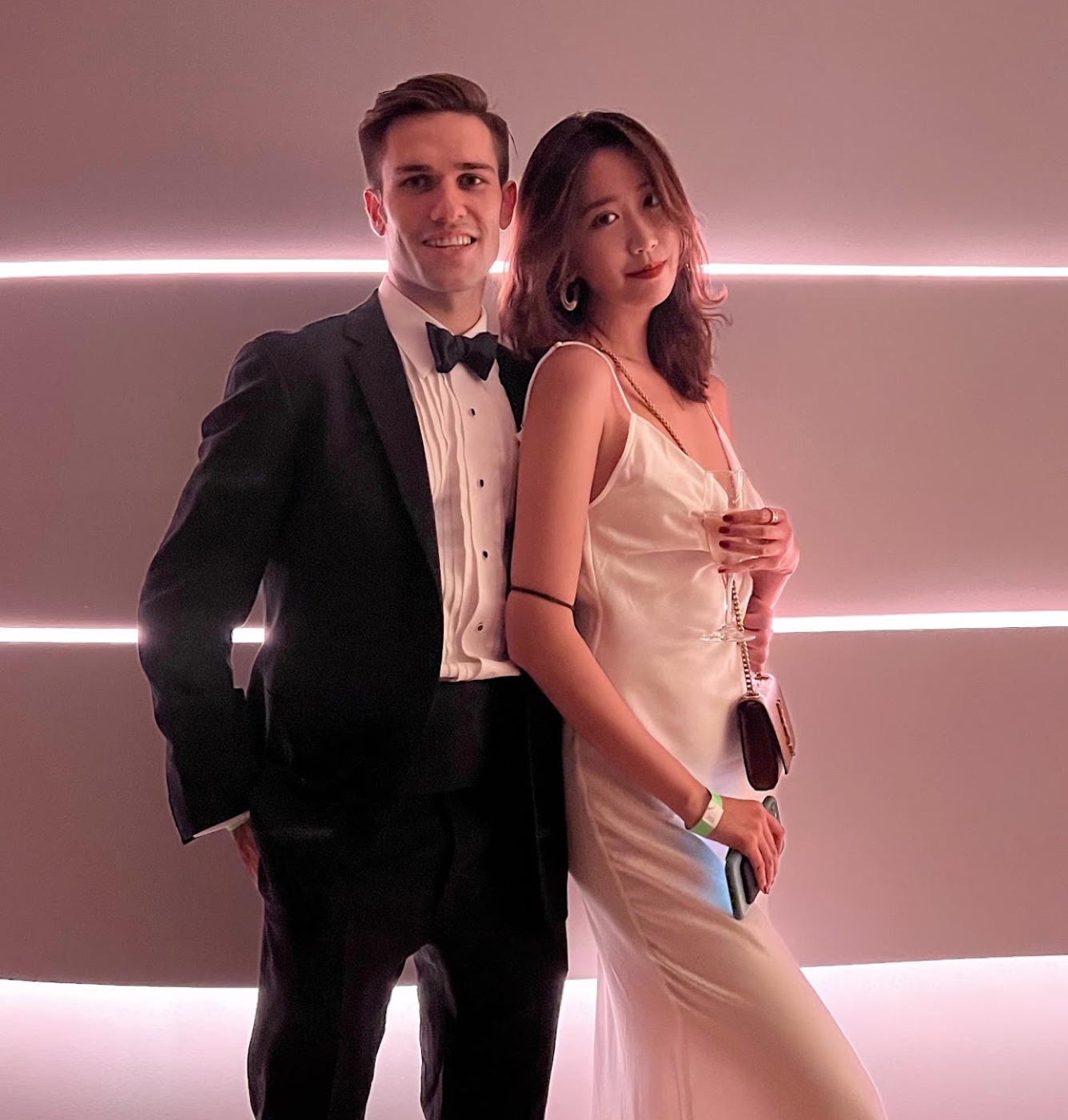 Noel in a tuxedo and a woman in a white slip dress with a small YSL bag pose back to back against a pink-lit sculpted wall.