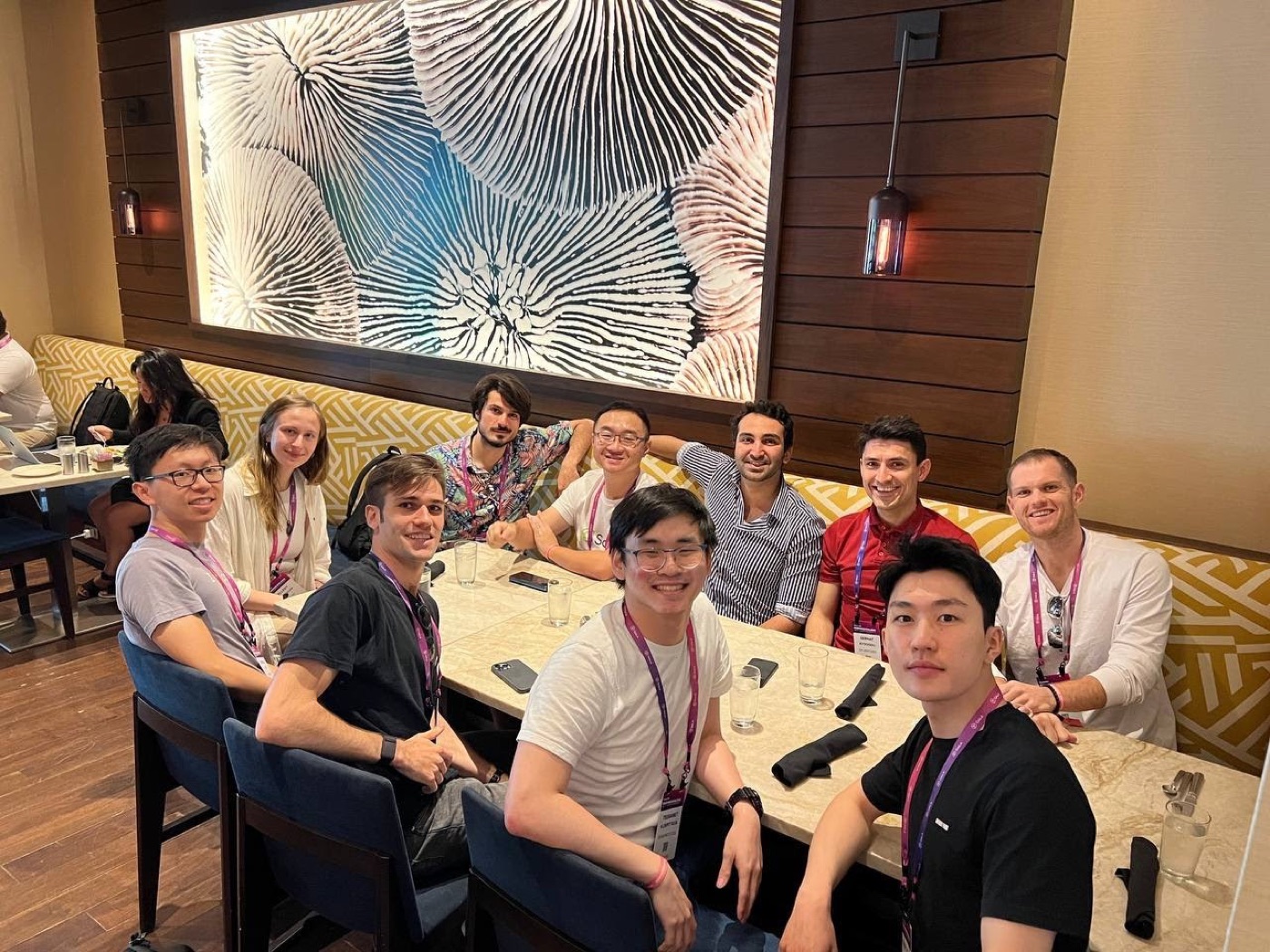 Ten conference attendees in lanyards share a long marble restaurant table beneath a large coral-print artwork.