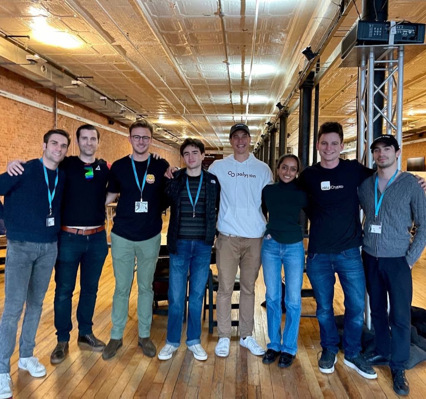 Eight people link arms for a group photo in a tin-ceilinged brick loft, wearing event lanyards, a Polygon hoodie, and an a16z Crypto tee.