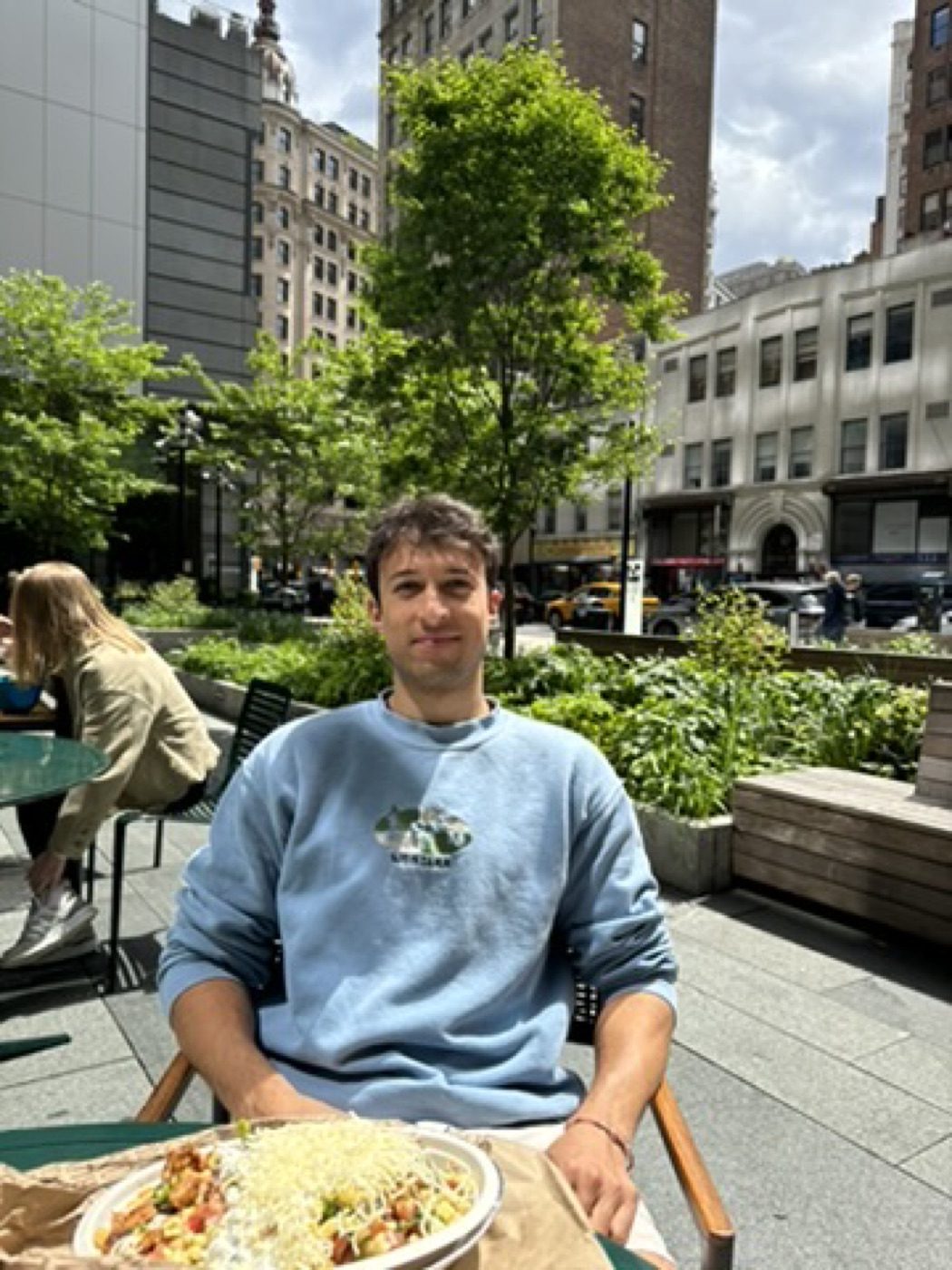 Man in blue sweatshirt at a leafy outdoor city cafe