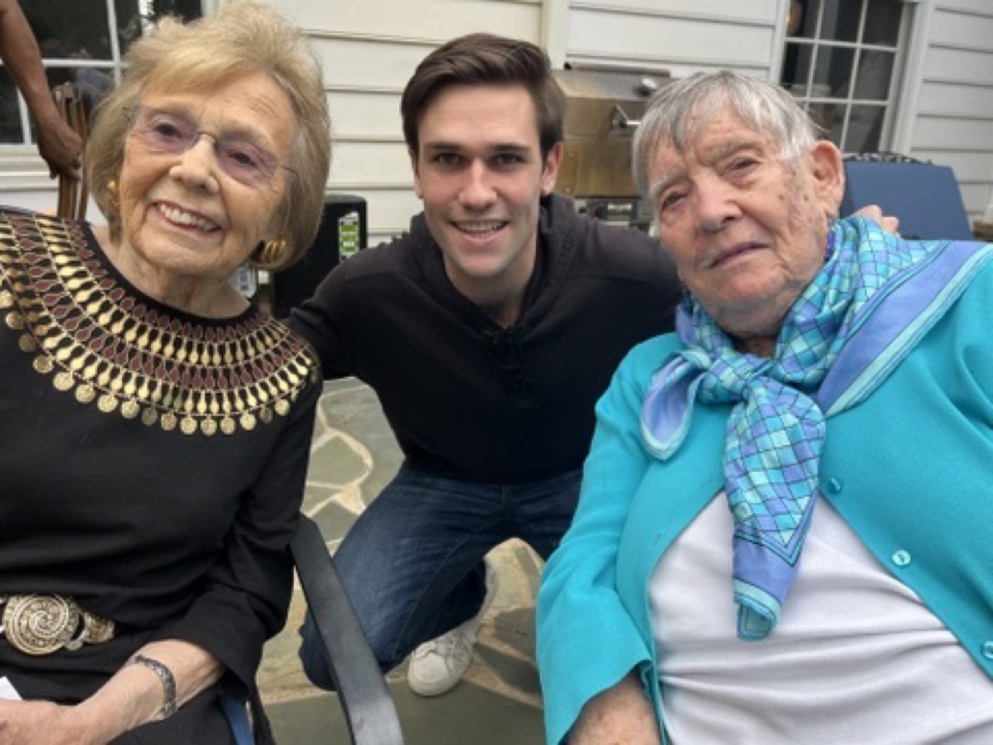 Noel beaming between his two grandmothers on a sunny patio
