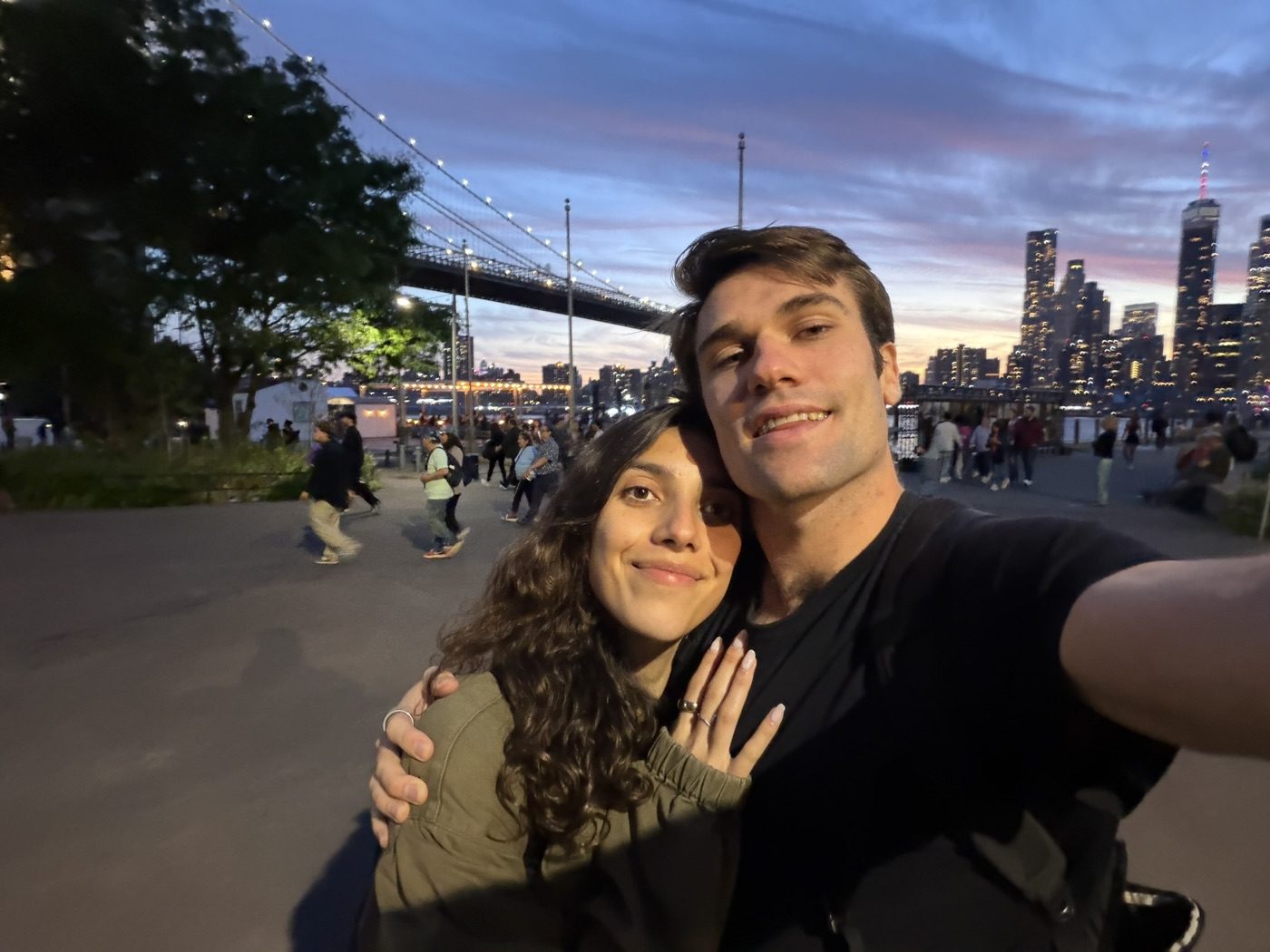 Cheek-to-cheek couple selfie under the Brooklyn Bridge at dusk