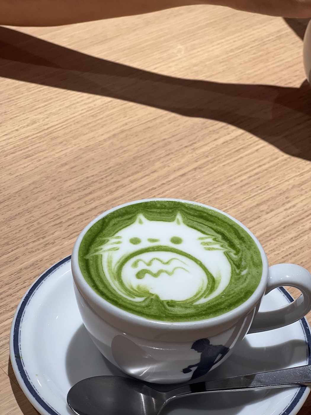 A matcha latte with Totoro drawn in the foam sits on a saucer on a wooden table.