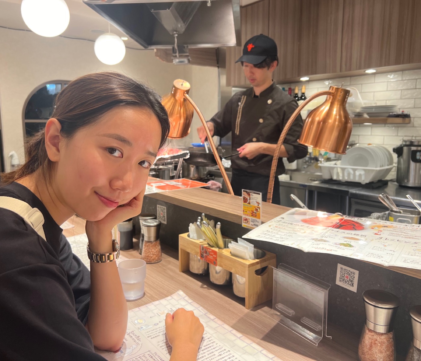 A woman rests her chin in her hand at a Japanese restaurant counter while a chef in a black cap cooks under copper heat lamps.