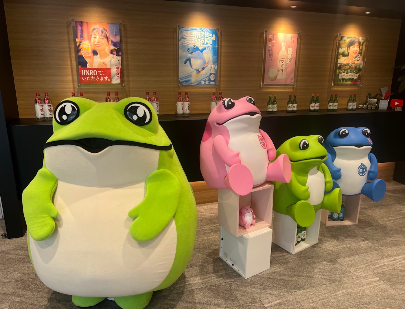 Four giant frog mascots in green, pink, and blue pose in a Jinro soju showroom lined with posters and bottles.